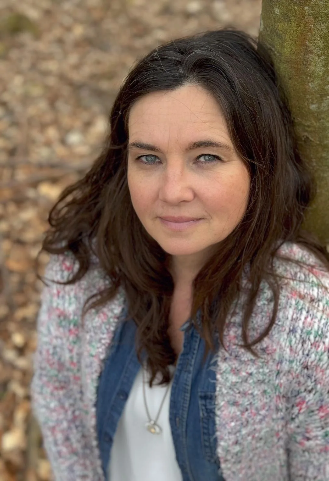 Yvonne Gröger lächelt leicht, dunkle lange Haare, Strickjacke mit Jeansweste, vor einem Baumstamm