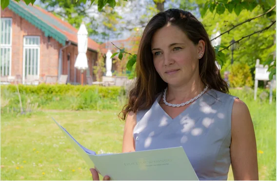 Yvonne Gröger mit der Mappe ihrer Traurede in einem Garten, im Hintergrund ein Backsteinhaus